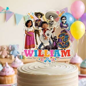 May include: A white frosted cake with a colorful Coco-themed cake topper that reads "WILLIAM" and the number "3". The topper features cartoon characters, musical notes, and a decorative border. Balloons in pink, blue, yellow, and purple are in the background.