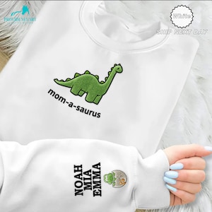 May include: White sweatshirt with a green embroidered dinosaur and the text "mom-a-saurus". The sleeve features embroidered names and a small graphic. The sweatshirt is a crew neck style.