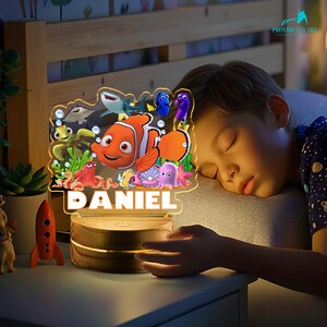 May include: A personalised night light with characters from "Finding Nemo." The lamp has a wooden base and a clear acrylic top with the name "DANIEL" illuminated. The design includes Nemo, Dory, and other sea creatures.