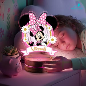May include: A Minnie Mouse themed night light in pink and black, featuring a bow, daisies, and the name "Victoria". The light sits on a wooden base. A pink piggy bank is nearby.