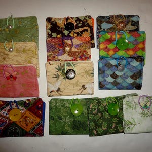 May include: A collection of handmade fabric wallets in various colors and patterns. Each wallet features a unique design, including floral, geometric, and abstract prints. They are secured with elastic loops and decorative buttons, offering a charming and functional accessory.