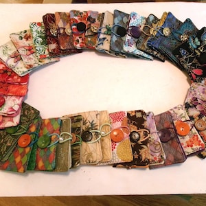 May include: A collection of rectangular fabric pouches arranged in an oval shape. Each pouch features a unique patterned textile design, including floral, geometric, and abstract motifs, and is secured with a single button and elastic loop closure.