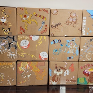 May include: A collection of twelve cardboard boxes, each hand-painted with colorful illustrations of Pokémon characters. The boxes feature various Pokémon, including Raichu, Golem, and Articuno, with their names and related text.