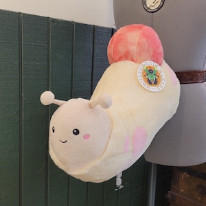 May include: A plush, smiling snail toy in cream and pink. The snail has a light pink head with small antennae and rosy cheeks. Its body is cream-colored with pink polka dots. A round, pink and orange shell sits on its back. A round sticker is attached to the snail.