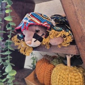 Puede incluir: Un animal de peluche con un sombrero estampado colorido y un parche negro en un ojo, descansando sobre una estantería de madera. Dos calabazas de ganchillo en naranja y amarillo se encuentran en una bandeja de madera debajo, con follaje verde a la izquierda.