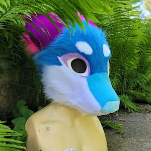 Op de afbeelding: Een kleurrijke fursuit-kop met een blauw, wit en turquoise ontwerp met roze en paarse accenten. De kop is op een mannequin gemonteerd en omgeven door groene bladeren. De ogen zijn groot en zwart.
