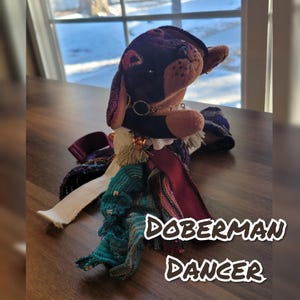 May include: A handmade plush Doberman dog with a burgundy and black head, tan snout, and a decorative collar. The dog is adorned with a white collar, tassels, and a scarf with the text "Doberman Dancer".