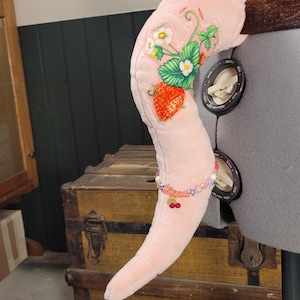 May include: A plush, light pink, curved tail accessory attached to a gray mannequin. The tail features embroidered strawberry plants with green leaves, white flowers, and red berries. A beaded bracelet with small cherry charms encircles the tail.