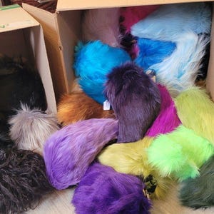 May include: A collection of colorful faux fur tails in various shades, including black, gray, blue, orange, purple, pink, green, and yellow, spilling out of cardboard boxes. The tails have a fluffy, textured appearance.