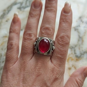 Könnte beinhalten: Ein silberfarbener Ring mit einem großen, ovalen, roten Edelstein. Der Ring hat ein rechteckiges Band mit dekorativen Details. Der Edelstein ist der Mittelpunkt, gefasst in einer detaillierten Lünette.