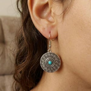 Könnte beinhalten: Silberfarbene, runde Ohrringe mit einem türkisfarbenen Mittelstein. Die Ohrringe haben ein detailliertes, kreisförmiges Design mit einer Reihe kleiner, sich wiederholender Muster an den Rändern. Die Ohrringe sind an einem einfachen Haken befestigt.