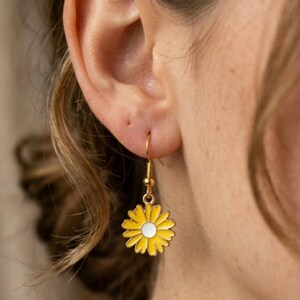 Könnte beinhalten: Ein goldfarbener Ohrring in Form einer Gänseblume mit gelbem Emaille-Blütenblatt-Design und weißem Zentrum. Der Ohrring ist ein Tropfenstil, der an einem goldfarbenen Haken hängt. Der Ohrring wird am Ohr einer Person gezeigt.