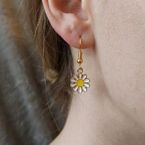 Könnte beinhalten: Goldfarbener Ohrring mit einer Gänseblümchen-Blume aus weißem und gelbem Emaille. Der Ohrring ist im Tropfenstil mit einem Hakenverschluss. Das Gänseblümchen hat ein detailliertes Design.