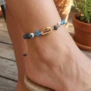 Könnte beinhalten: Fußkettchen mit einem goldenen Muschelanhänger, blauen Perlen und kleinen Evil-Eye-Perlen. Das Armband ist an einer dünnen, beigefarbenen Kordel befestigt. Das Fußkettchen wird an einem Fuß mit einer neutralfarbenen Sandale getragen.
