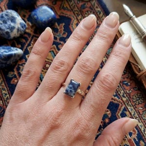 Könnte beinhalten: Ein silberner Ring mit einem rechteckigen blauen Edelstein mit weißen Flecken. Der Ring befindet sich an einer Hand, im Hintergrund ein gemusterter Teppich und blaue Steine. Weitere Elemente sind eine Lupe.