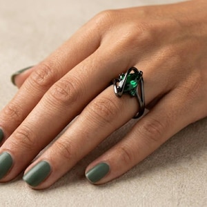 Könnte beinhalten: Ein schwarzer Ring mit einem großen, smaragdgrünen Edelstein und kleineren grünen Akzenten. Der Ring hat ein einzigartiges, wirbelndes Design. Die Hand, die den Ring trägt, hat grünen Nagellack.