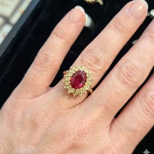 Könnte beinhalten: Ein goldener Ring mit einem großen, oval geschliffenen Rubin. Der Rubin ist von einem Kranz aus kleineren, runden Edelsteinen umgeben. Der Ring ist auf einem goldenen Band gefasst. Der Ring befindet sich an einem Finger.