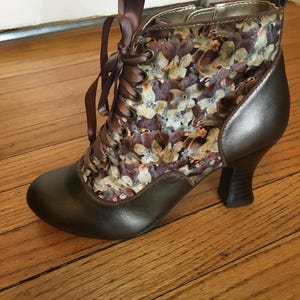 May include: Brown ankle boot with a floral patterned fabric upper and a brown leather toe. The boot features a lace-up design with brown ribbon laces and a small heel. The boot has a metallic bronze accent on the heel.