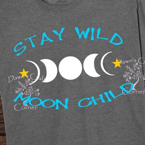 Puede incluir: Una camiseta gris con el texto "Stay Wild Moon Child" en azul y amarillo. El dise&ntilde;o incluye tres lunas y estrellas.