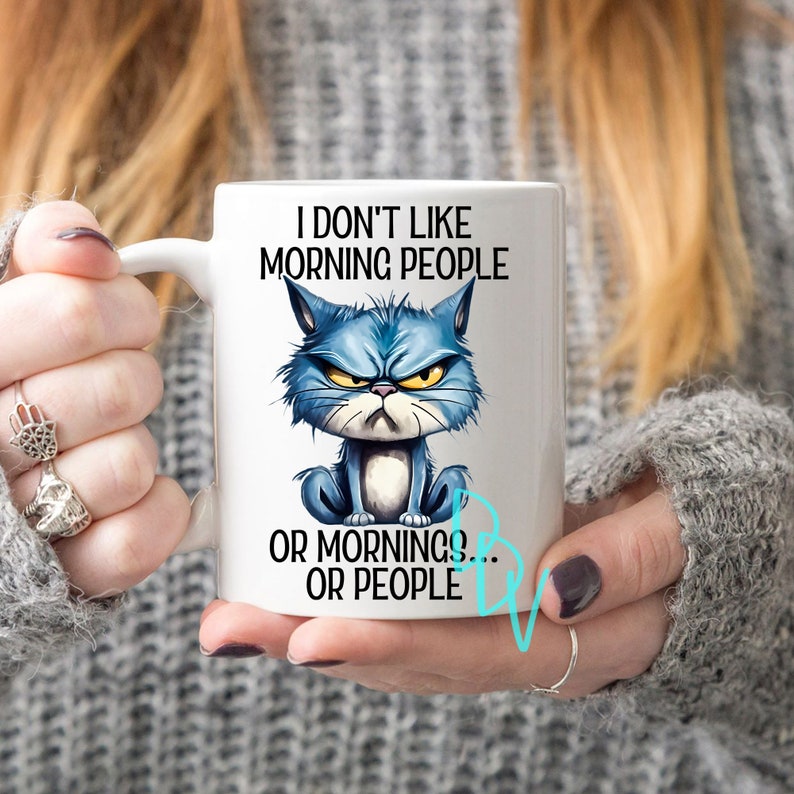 May include: White ceramic mug with the text "I DON'T LIKE MORNING PEOPLE OR MORNINGS... OR PEOPLE" and a grumpy blue cat illustration. The mug has a white handle and is held by a person.