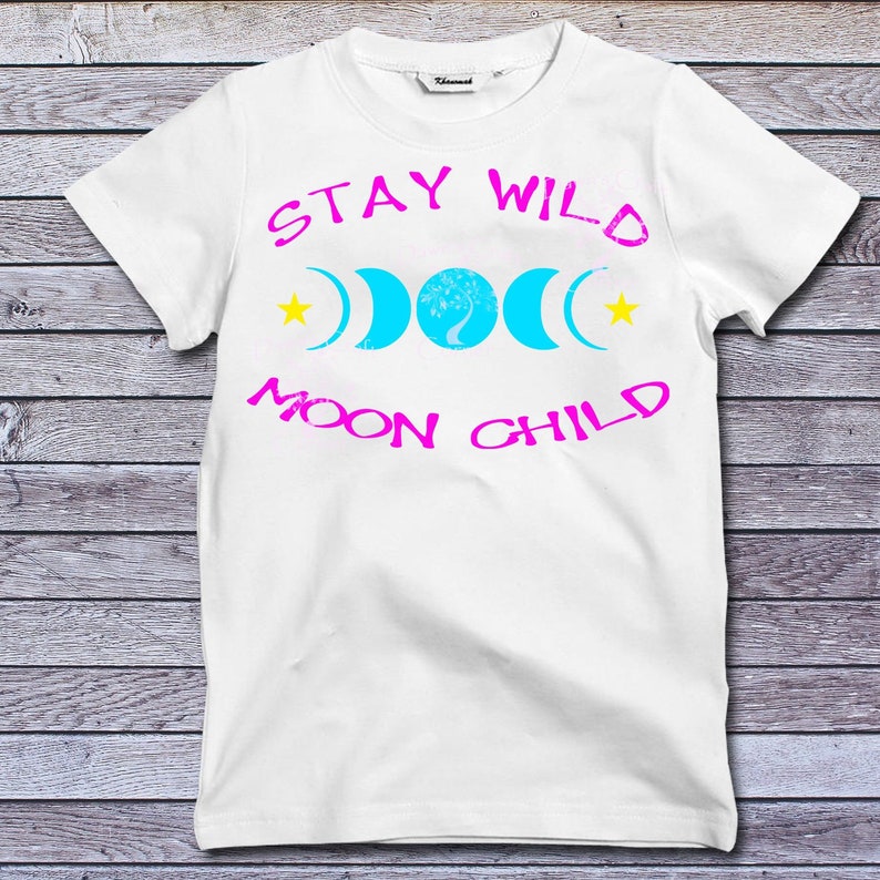 Puede incluir: Camiseta blanca con texto rosa que dice "Stay Wild Moon Child". El dise&ntilde;o tambi&eacute;n incluye una luna creciente azul con un &aacute;rbol en su interior y dos estrellas amarillas.