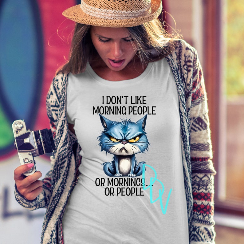 May include: White t-shirt with the text "I DON'T LIKE MORNING PEOPLE OR MORNINGS... OR PEOPLE." The shirt features a cartoon illustration of a grumpy blue cat with yellow eyes. The cat is sitting with its paws crossed.