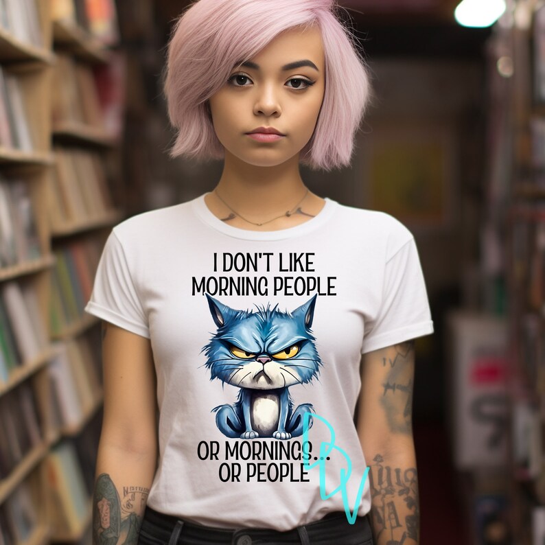 May include: White t-shirt with the text "I don't like morning people or mornings... or people." A blue cartoon cat with an angry expression is featured below the text. The shirt is being worn by a person with pink hair.