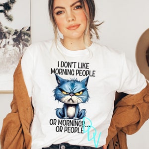 May include: White t-shirt featuring a grumpy blue cat illustration with the text "I DON'T LIKE MORNING PEOPLE OR MORNINGS... OR PEOPLE". The cat has an angry expression with yellow eyes. The shirt is a casual wear item.
