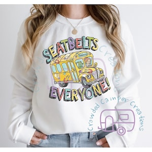May include: White sweatshirt featuring a yellow school bus graphic with the text "SEATBELTS EVERYONE!" in colourful lettering. The bus has a green monster and a girl with red hair inside. The text "Crowded Camper Creations" is written in purple in the bottom right corner of the sweatshirt.