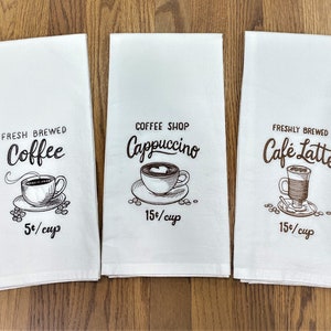 May include: Three white kitchen towels with brown text and illustrations of coffee, cappuccino, and cafe latte. The text reads "Fresh Brewed Coffee 5¢/cup", "Coffee Shop Cappuccino 15¢/cup", and "Freshly Brewed Cafe Latte 15¢/cup".