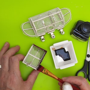 Może przedstawiać: Zbliżenie na osobę malującą białą siatkową klatkę czarną farbą. Klatka jest wykonana z drutu i ma prostokątny kształt. Osoba używa pędzla do nakładania farby. Na stole znajduje się mała miseczka z czarną farbą i nożyczki.