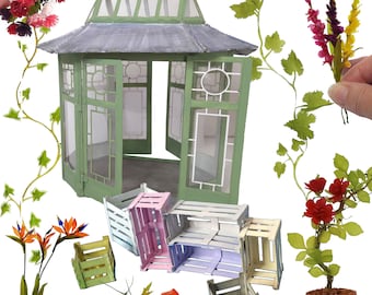 Puede incluir: Un invernadero en miniatura verde y blanco con un techo gris y puertas abiertas. El invernadero está rodeado de varias plantas y flores en miniatura, incluyendo un rosal en maceta, un arreglo floral en maceta y una planta de hiedra en maceta. También hay varias cajas de madera en miniatura de varios colores.