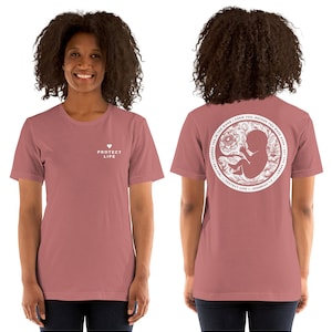 Peut inclure: Un t-shirt rose avec un cœur blanc et le texte "PROTECT LIFE" sur le devant. Le dos du t-shirt présente un cercle blanc avec un motif floral et un fœtus à l'intérieur. Le texte "I FORMED YOU IN THE WOMB, I KNEW YOU BEFORE YOU WERE BORN, I SET YOU APART FOR ME" est écrit autour du cercle. Le texte "PROTECT LIFE - JEREMIAH 1:5" est écrit sous le cercle.