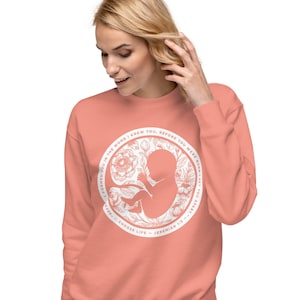 Puede incluir: Sudadera de color coral con un diseño circular blanco. El diseño incluye un estampado floral y una silueta fetal. El texto dice: "TE FORME EN EL VIENTRE, TE CONOCÍ, ANTES DE QUE NACIERAS, TE APARTE - ELIGE LA VIDA - JEREMÍAS 1:5."