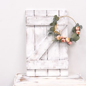 May include: A whitewashed wooden door with a gold metal wreath adorned with pink flowers and green leaves. The wreath has the word "hi" written in gold script.