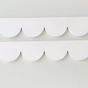 May include: Two white, scalloped-edge decorative trim pieces. The trim has a smooth, matte finish and a repeating pattern of rounded curves. The pieces are positioned horizontally against a light-colored background.