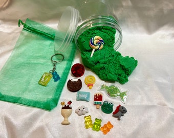 Puede incluir: Un tarro verde lleno de arena cinética verde, una bolsa de cordón verde, un llavero con un amuleto de osito de goma y un amuleto de barra de caramelo, y varios amuletos coloridos que incluyen una piruleta, un osito de goma, una rana, un dinosaurio, una paleta, una nube, una magdalena y una barra de caramelo.