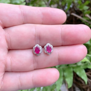 Könnte beinhalten: Ein Paar silberne Ohrstecker mit einem roten ovalen Rubin, der von einem Heiligenschein aus kleineren weißen Edelsteinen umgeben ist.