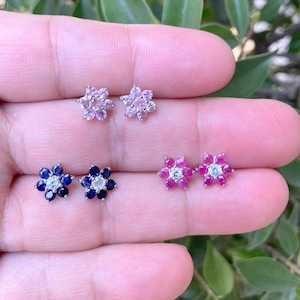May include: Four pairs of flower-shaped stud earrings with different colored gemstones. The earrings are silver and have a white center stone surrounded by smaller colored stones. The colors are pink, blue, and red.