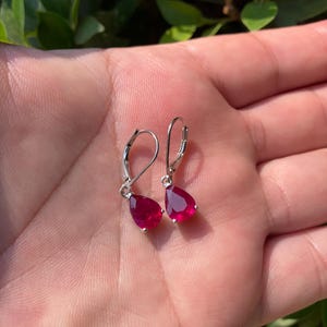 May include: A pair of silver-toned drop earrings with teardrop-shaped, deep red gemstones. The earrings are displayed on a hand, with a blurred green background.