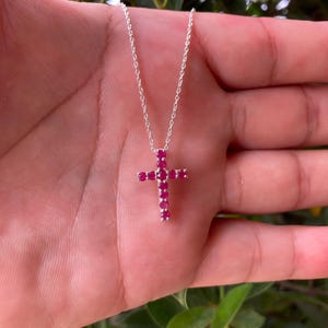 Puede incluir: Un collar de cadena de plata con un colgante de cruz engastado con pequeñas piedras preciosas redondas de color rojo rosado vibrante. La cruz está formada por una fila vertical de cinco piedras y una fila horizontal de tres piedras.