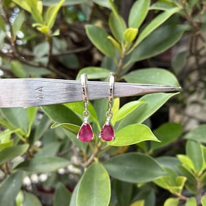 May include: A pair of silver-toned hoop earrings featuring dangling teardrop-shaped pink gemstones. The earrings are held by a pair of metal tweezers marked with the letters AA, showcasing the small size of the jewelry pieces.