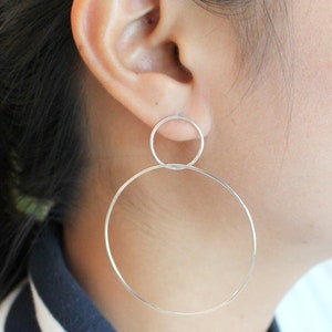 May include: Silver hoop earrings. The earrings feature a small circle connected to a larger hoop. The earrings are worn on the ear, with the ear and part of the neck visible in the image. The earrings are a simple, elegant design.