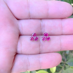 May include: Two small stud earrings, each with three vibrant, round, ruby-coloured gemstones. The earrings are set in a silver-coloured metal, forming a delicate floral design. The background is a soft, blurred green.