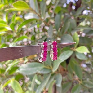 May include: A pair of silver-toned hoop earrings, each featuring a vertical row of four round, vibrant pink gemstones. The earrings are held by a pair of metal tweezers, showcasing the polished metal setting and the faceted texture of the stones.