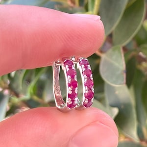 Könnte beinhalten: Ein Paar silberne Creolen, jede mit einer Reihe leuchtend roter Edelsteine besetzt. Die Ohrringe werden zwischen Fingern gehalten, wodurch das Design und die Farbe des Schmucks vor einem Hintergrund aus grünen Blättern zur Geltung kommen.