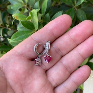 Puede incluir: Un par de pendientes de aro plateados con colgantes en forma de corazón y cuadrado de color rojo rubí. Los pendientes se muestran sobre un fondo de hojas verdes.