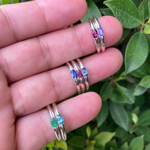 May include: A collection of seven silver rings, each featuring a single small oval-cut gemstone in various colors including pink, blue, red, and green. The rings are arranged in groups on fingers, showcasing polished silver bands and vibrant, faceted stones.
