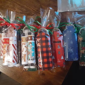 May include: A collection of 10 small, clear plastic bags filled with lip balm tubes. The lip balms are wrapped in festive holiday designs, including plaid, snowflakes, and Christmas trees. The bags are tied with red and green ribbon.