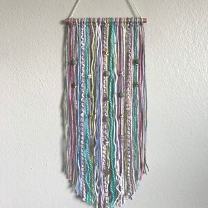 May include: A handmade wall hanging featuring vertical strands of yarn in various colors, including gray, pink, blue, and yellow. The yarn is adorned with decorative beads and star-shaped charms. The wall hanging is suspended from a wooden dowel with a white string.